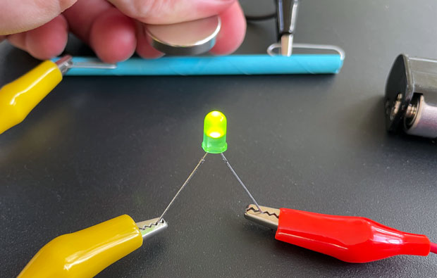 a green LED is lit up. In the background, someone holds a magnet near a straw.
