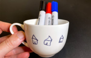 A tecup with 3 houses drawn on it. there are 3 different whiteboard markers inside the cup
