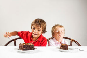 Two kids, one with a big slice of cake and one with a smaller slice.