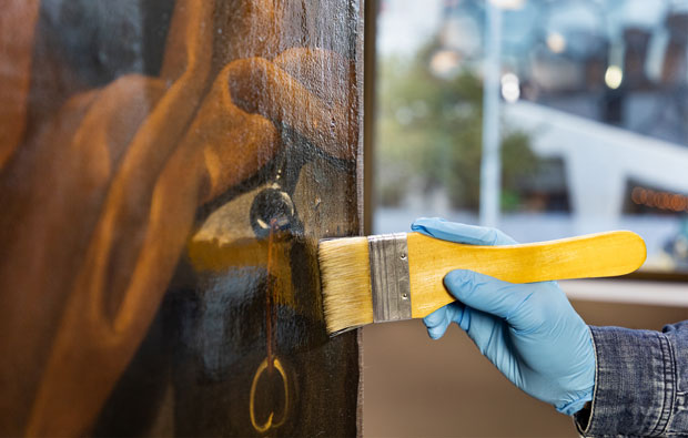 Gloved hand painting varnish with a brush onto a work of art.