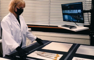 Person in labcoat, mask and gloves looking at a screen and pressed plant specimens