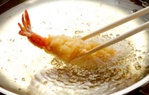 A prawn held in chopsticks, sizzling in a pan.